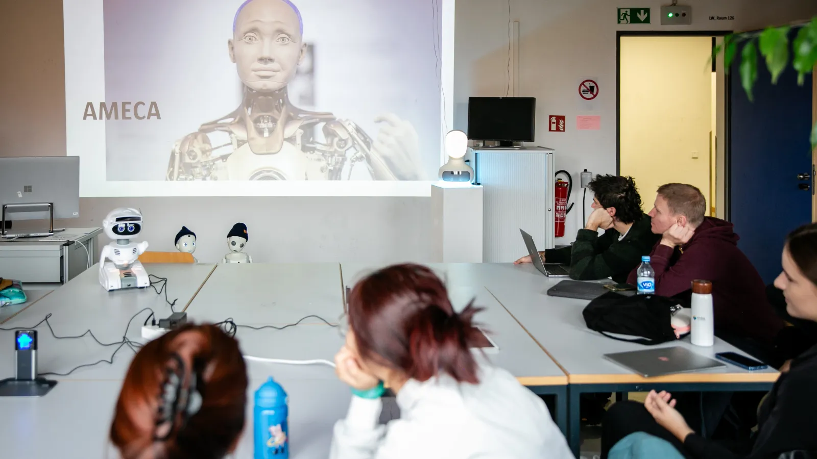 Studierende sitzen im Social Robotic Lab um einen Tisch herum, auf dem verschiedene kleine Roboter stehen; an die Wand wird ein Bild des Roboters Ameca projiziert