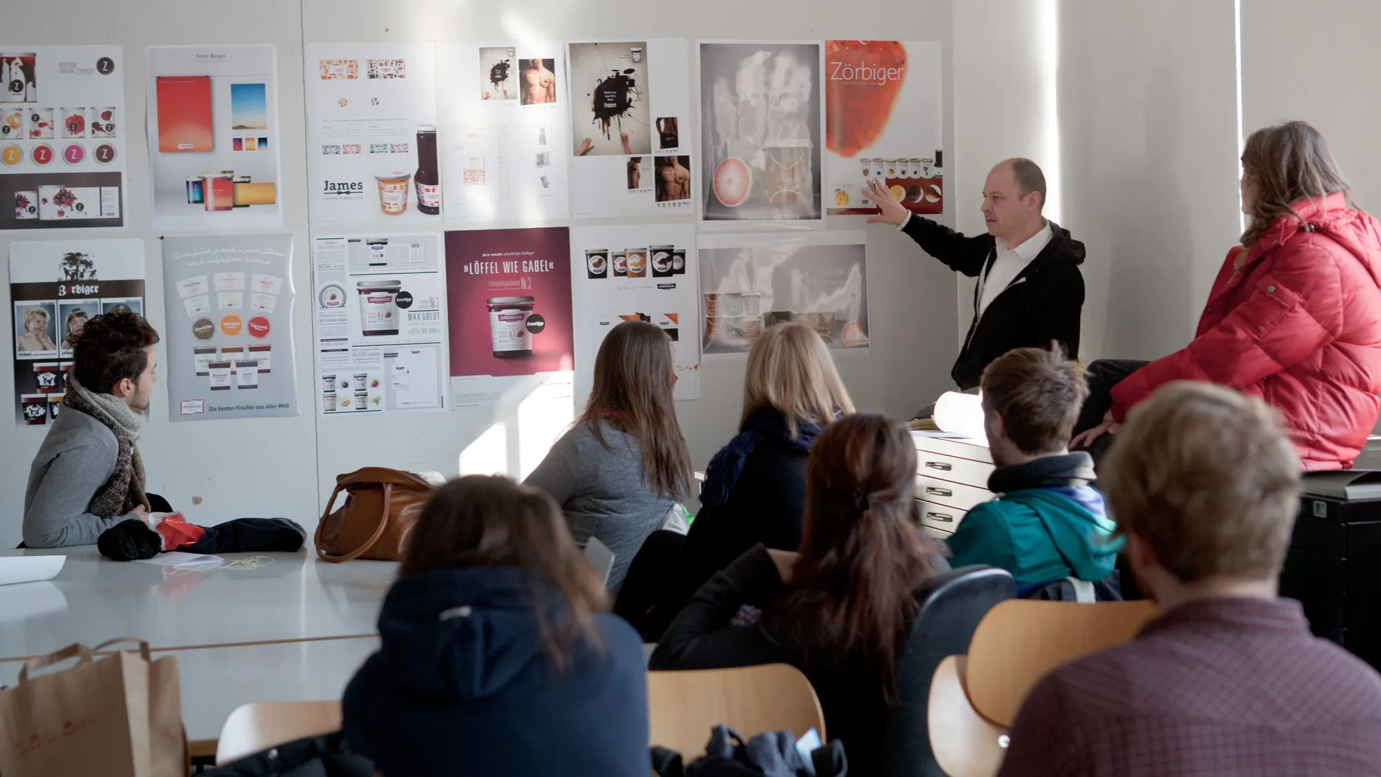 Lehrender zeigt vor Studierenden auf Entwrfe an einer Wand aus dem Studiengang Kommunikationsdesign