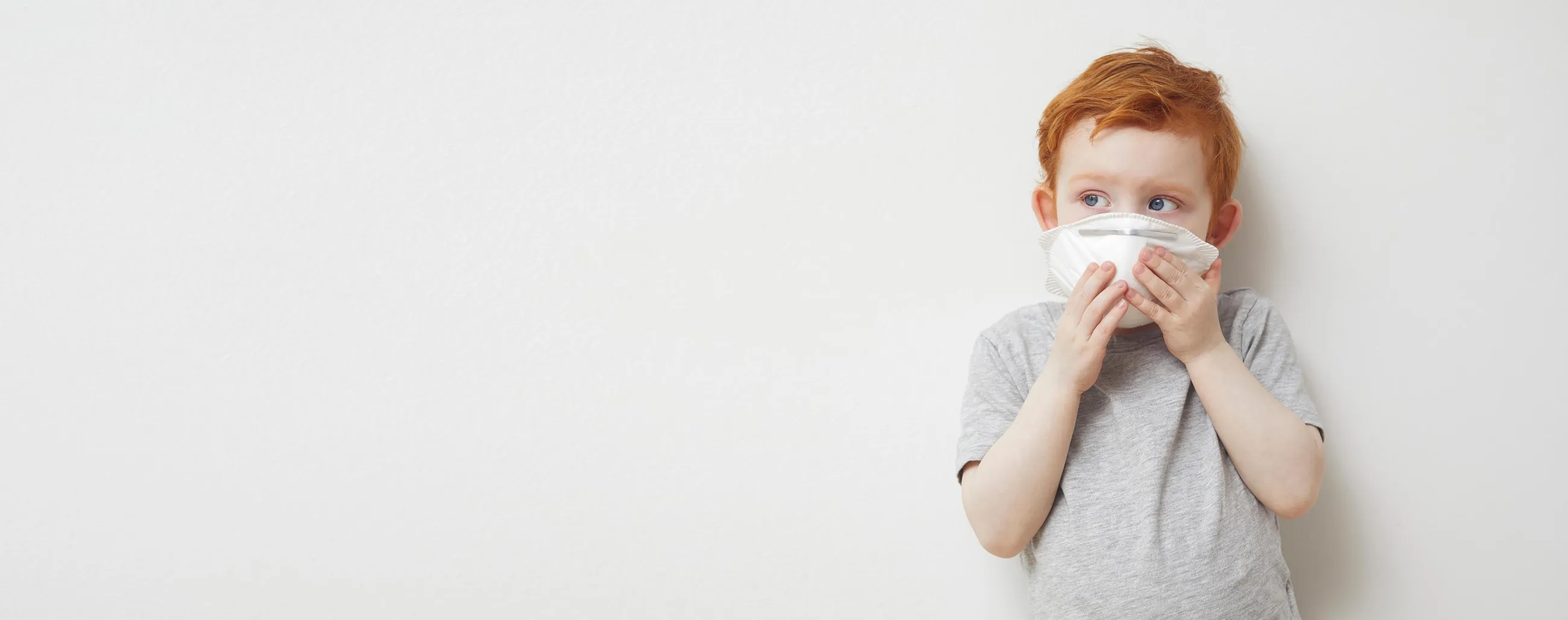 Junge mit Corona-Maske vor dem Gesicht, steht vor heller Wand